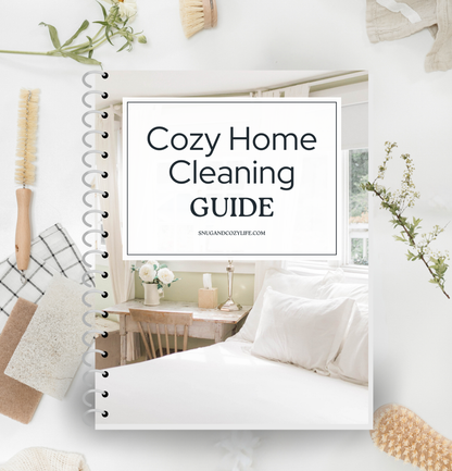 this is a mockup of our cozy home cleaning guide on a binder. there are cleaning brushes, sponges, rags, and sprigs of flowers surrounding it on the table.