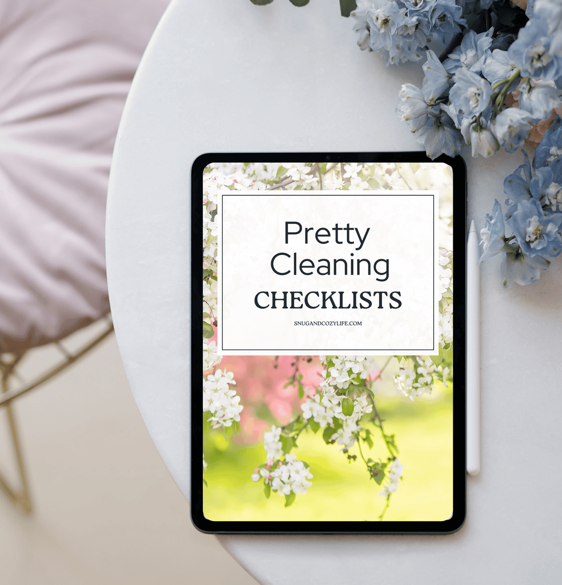 A tablet and purple flowers sit on a round white table. The table shows a white-flowered tree with the words &quot;Pretty Cleaning Checklists&quot; in a text box above it.